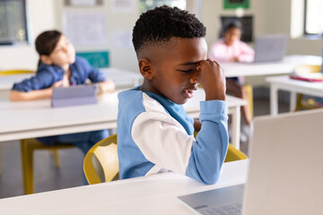 Diverse child classmates in classroom, African American boy in blue top using laptop, tablet nearby