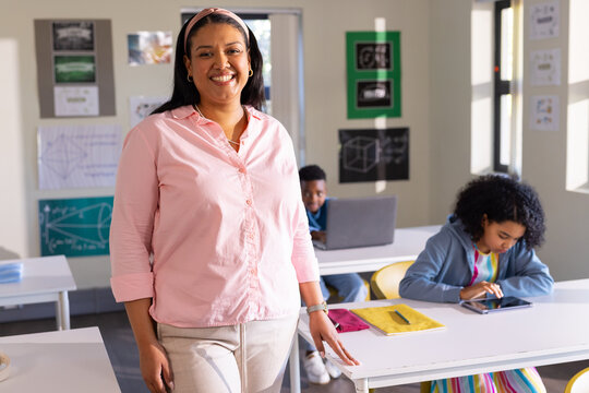 African American teacher standing smiling at camera in class with students using tablet and laptop
