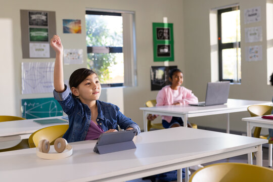 Diverse children in classroom sitting at desks raising hands while using tablet, laptop, headphones