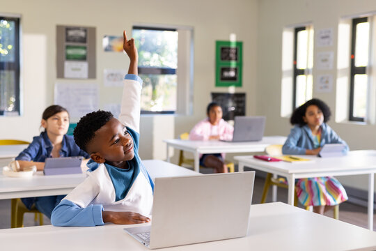 Diverse classmates with African American boy raising hand using silver laptop in classroom