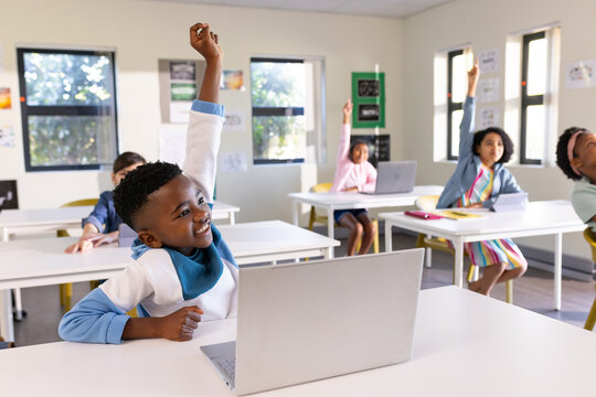 Diverse children sitting in classroom using laptops and raising hands at white desks with posters