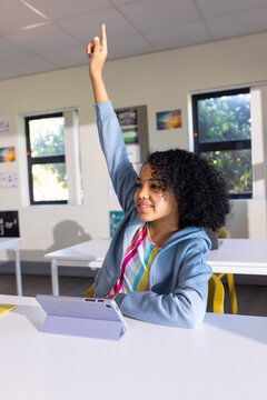 African American teen student sitting at white desk raising hand using purple tablet in classroom