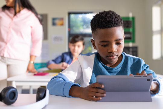 Students with teacher guiding African American boy using tablet on desk, headphones nearby