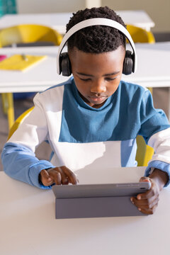 African American boy using tablet in folio case at school desk, wearing white headphones