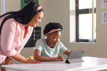 African American woman leaning over classroom table guiding girl tapping tablet folio case