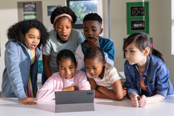 Diverse youth students leaning over tablet at classroom table using dark gray folio case, focusing
