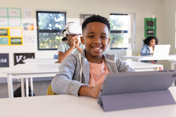 Diverse students in classroom, male in grey-hoodie smiling using tablet folio, VR headset, laptop