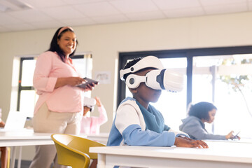 Teacher leading school pupils, boy sitting with white VR set, teacher holding tablet in classroom