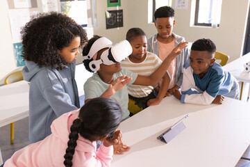 African American preteens gathering at school watching child wearing white VR headset using tablet