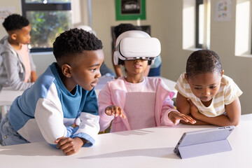 African American children using white VR headset, tablet in class at table center in pink jacket