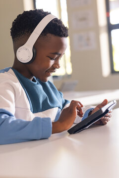 African American teen sitting at white desk in classroom wearing white headset using black tablet