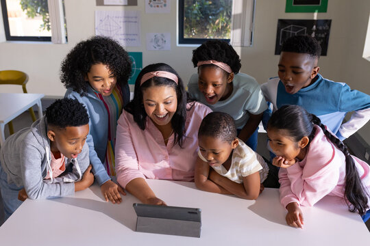 African American teacher with six youths leaning around tablet, operating device at classroom table