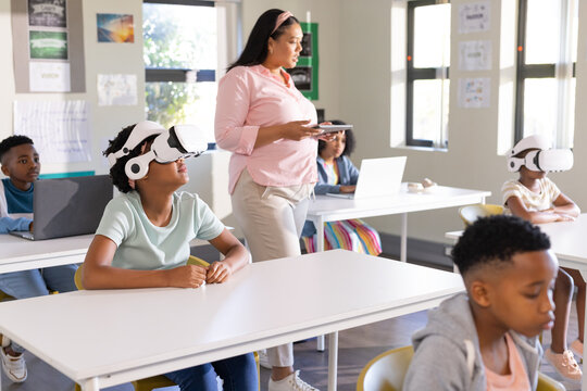 African American teacher holding tablet supervising students using VRs and laptops in classroom