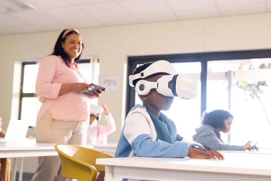Teacher leading school pupils, boy sitting with white VR set, teacher holding tablet in classroom