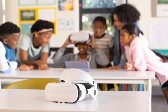 African American students watching classmate wearing VR and using tablet, white VR on desk in class