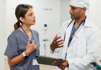 African American doctor and nurse discussing patient info while holding tablet in clinic corridor