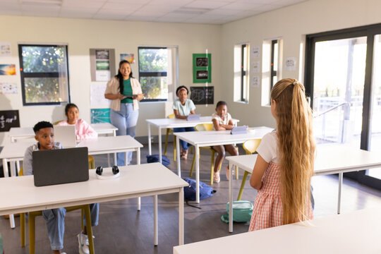 Diverse class watching girl presenting at front of class with laptop wearing pink dress, copy space