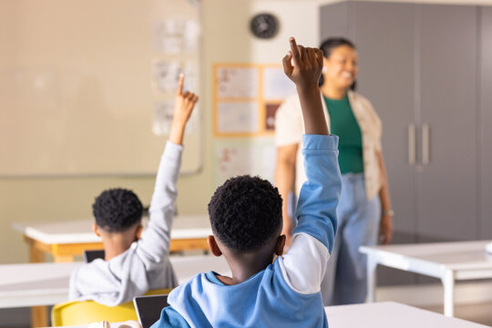 African American boy sitting at desk wearing blue sweatshirt, raising right arm toward whiteboard