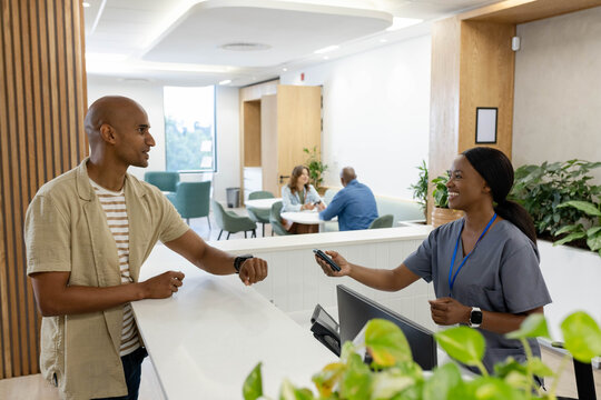 African American receptionist wearing grey scrubs holding terminal as visitor leaning at reception