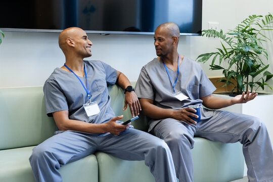 African American males in grey scrubs sitting and chatting on lounge bench, holding phone and mug