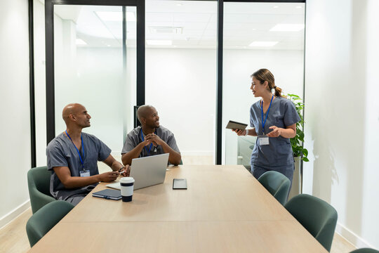 Medical team wearing grey scrubs meeting in clinic, woman presenting on tablet as team using laptop