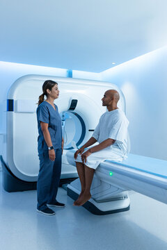 African American clinician navy-scrubs talking with patient sitting on CT table in gown near gantry