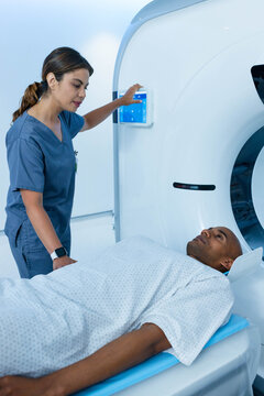 Female nurse wearing scrubs operating panel while male patient lying on CT table in gown