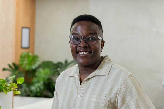 Adult man standing and smiling in office lounge wearing beige collared shirt, clear glasses, plants