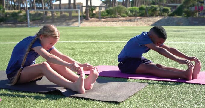 Diverse teenage classmates following coach cue, stretching forward on mats on turf near soccer goal