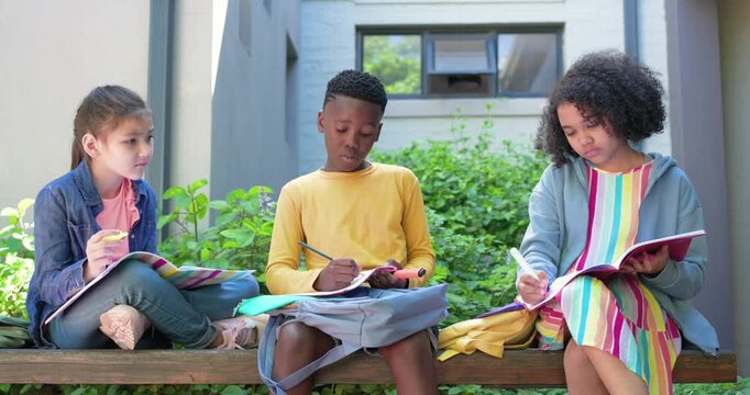 Diverse children writing on backpack with pencils at courtyard bench, prompted by open notebooks