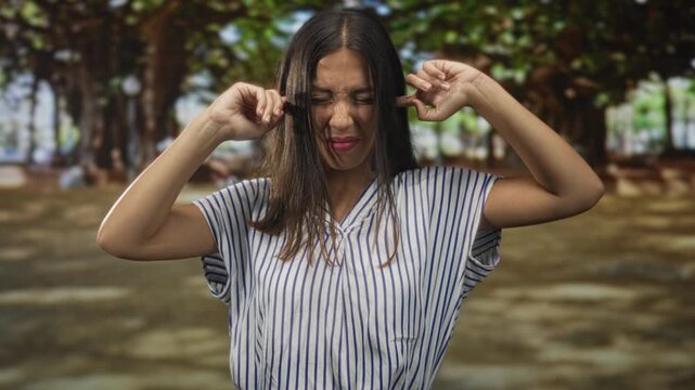 Hispanic young brunette woman plugs ears with fingers while grimacing in green park among large trees and dappled sunlight; irritation avoidance solitude.