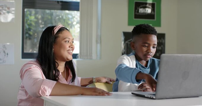 Asian woman leaning forward speaking and tutoring African American youth boy typing laptop at desk