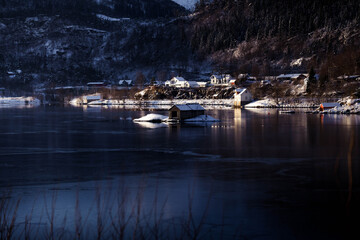 Fototapeta premium Small villages with red houses sit near the Norwegian fjords, where sunlight highlights the snow-covered mountains and brings warm colors to the dramatic winter landscape.