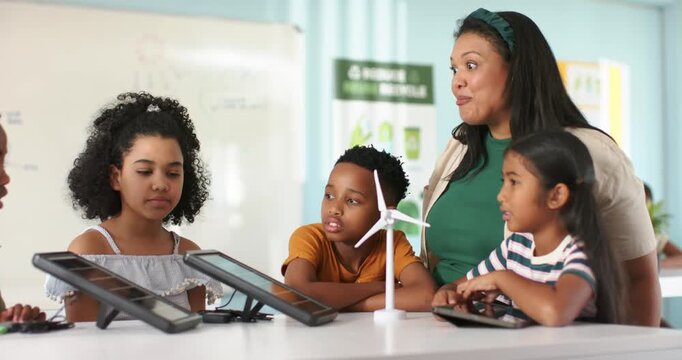 Teacher with teens watching offframe hand pointing at turbine and tablets at table, learning STEM