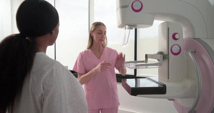 Rad tech in pink scrubs showing breast placement at mammogram to African-American patient listening