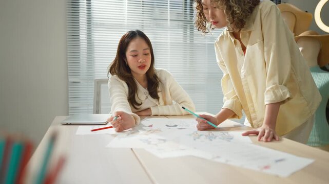 Two creative Asian fashion designers collaborate intensely in their bright studio workspace, brainstorming innovative ideas and sharing design concepts for their upcoming seasonal clothing collection