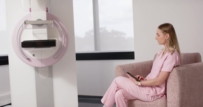 Adult woman in pink scrubs sitting in clinic tapping tablet, machine drawing glances, copy space