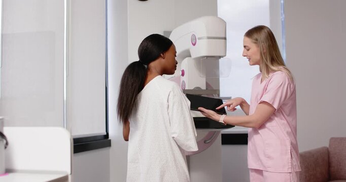 Diverse patient placing hand on mammogram; pink-scrub tech gesturing with tablet guiding for scan