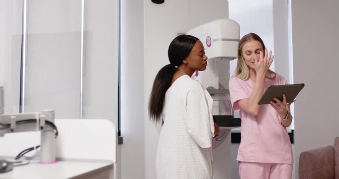 Diverse female patient white gown listening, tech tapping tablet explaining mammogram in exam room