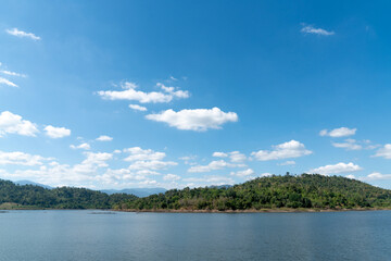 Scenic landscape of a calm blue lake surrounded by lush green forested hills under a bright blue sky with scattered white clouds. Peaceful natural outdoor environment in a sunny tropical region.