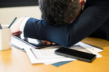 A middle-aged male professional in a suit resting his head on his arms over a digital tablet, showing signs of exhaustion and burnout in an office setting.