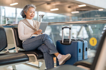 Happy senior asian woman using digital tablet at airport terminal while waiting for flight, elderly female tourist holding passport and booking online, retirement travel and technology concept