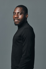 Obraz premium African man portrait in creative conceptual visual style wearing black turtleneck sweater on gray background. Confident young male with beard and short hair looking at camera.