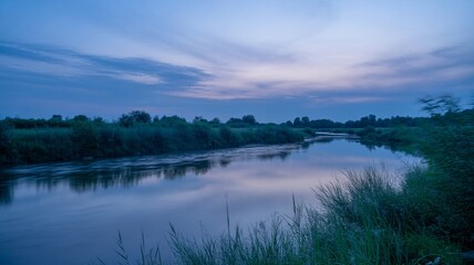 Fototapeta premium A beautiful sunset reflects over the calm water of a forest lake and river landscape as the sun glows through clouds in the evening sky