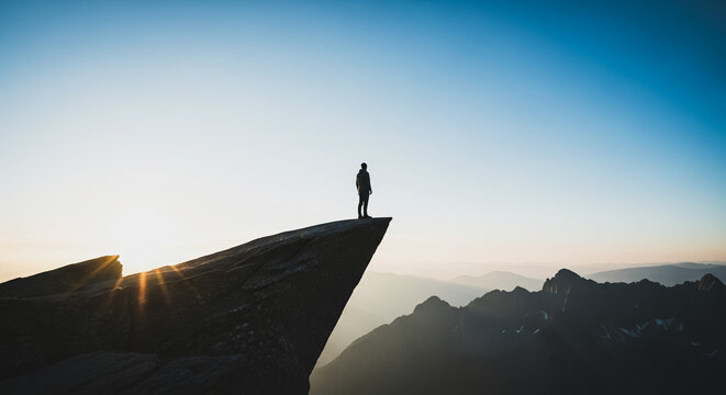 Silhouette of a man standing on the edge of a steep cliff overlooking mountain peaks at sunrise, symbolizing freedom, challenge, and success.
