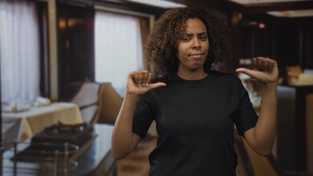 Young african american woman with thumbs pointing to self at a restaurant table inside a building; skepticism.