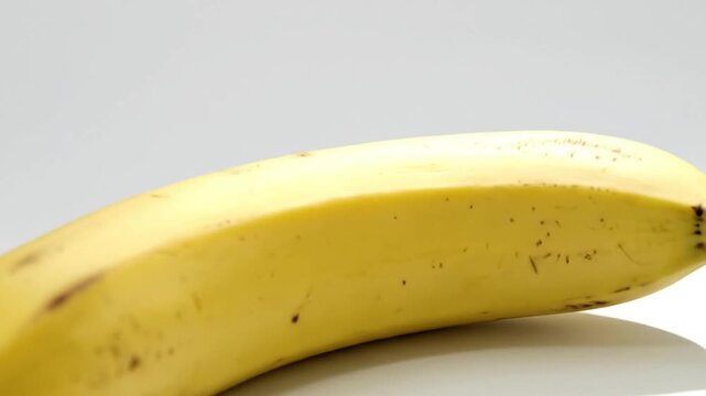 A close-up shot of a vibrant yellow ripe banana, beautifully isolated against a clean white background. The smooth skin of the fruit, adorned with subtle natural brown spots, is showcased in crisp det