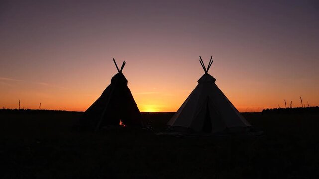 Two teepees silhouetted against a vibrant sunset with birds flying overhead.