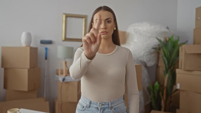 Young woman points finger at camera lens among stacked cardboard moving boxes, bubblewrap, lamp and plant in a room of a building; determination.