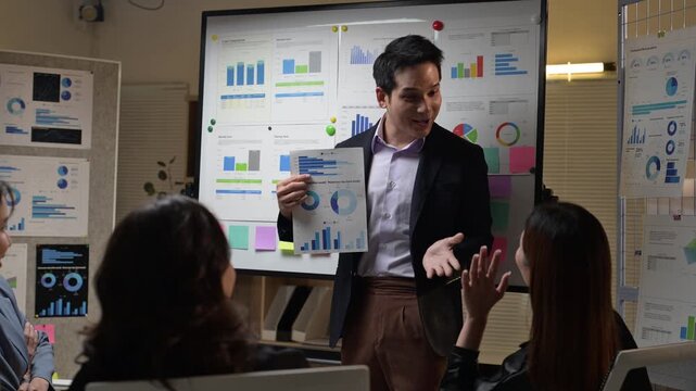 A man is standing in front of a large screen with a presentation. He is pointing to a piece of paper. There are three other people in the room, one of whom is sitting